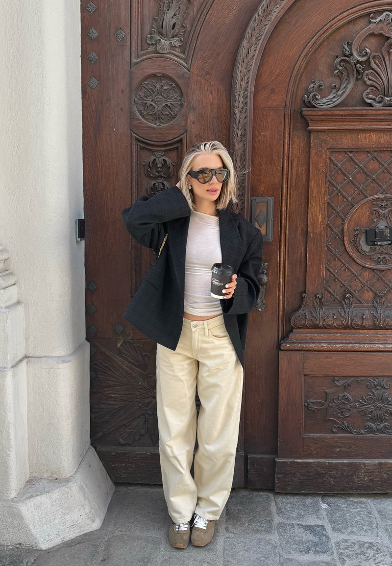 Blonde vrouw met oversized zwart blazer, beige broek en zonnebril, die een koffiekopje vasthoudt, staand voor een sierlijk gebeeldhouwde houten deur.