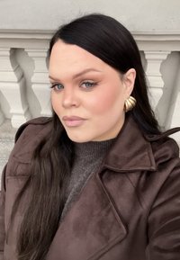 Bruine suède jas over een grijze trui, met lang zwart haar en gouden oorbellen in de vorm van schelpen, tegen een neutrale achtergrond.