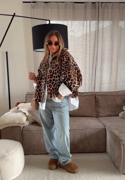 Chaqueta de piel sintética de estampado de leopardo sobre una camisa blanca, combinada con jeans holgados de color azul claro y botas de plataforma marrones hasta el tobillo. Entorno interior.