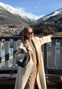 Femme en manteau beige et lunettes de soleil appuyée sur une rambarde en bois, contemplant des montagnes enneigées et un village sous un ciel bleu clair.