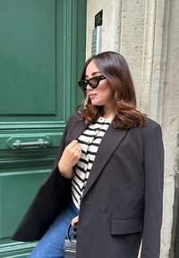 Übergroßer schwarzer Blazer über einem gestreiften Strickoberteil. Kombiniert mit blauen Jeans und schwarzen Sonnenbrillen. Schlichte Hardware und strukturierte Stoffdetails.