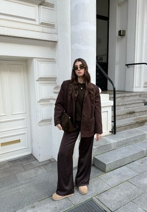 Mujer con blazer marrón oscuro, pantalones de pierna ancha, zapatos beige y gafas de sol, de pie en unas escaleras de piedra junto a la entrada de un edificio blanco.