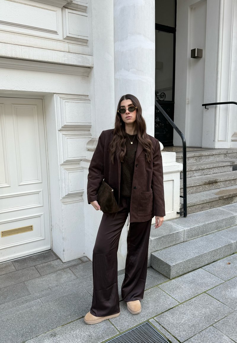 Donna con blazer marrone scuro, pantaloni a gamba larga, scarpe beige e occhiali da sole, in piedi su gradini di pietra vicino all'ingresso di un edificio bianco.