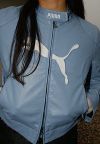 Veste en cuir bleu clair avec un logo Puma blanc bien visible sur la poitrine, col montant et poches zippées.