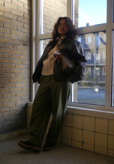 Chaqueta de cuero negra sobre un suéter blanco, pantalones de pierna ancha verde oliva, de pie junto a una ventana, sosteniendo un bolso marrón con acentos de llavero.