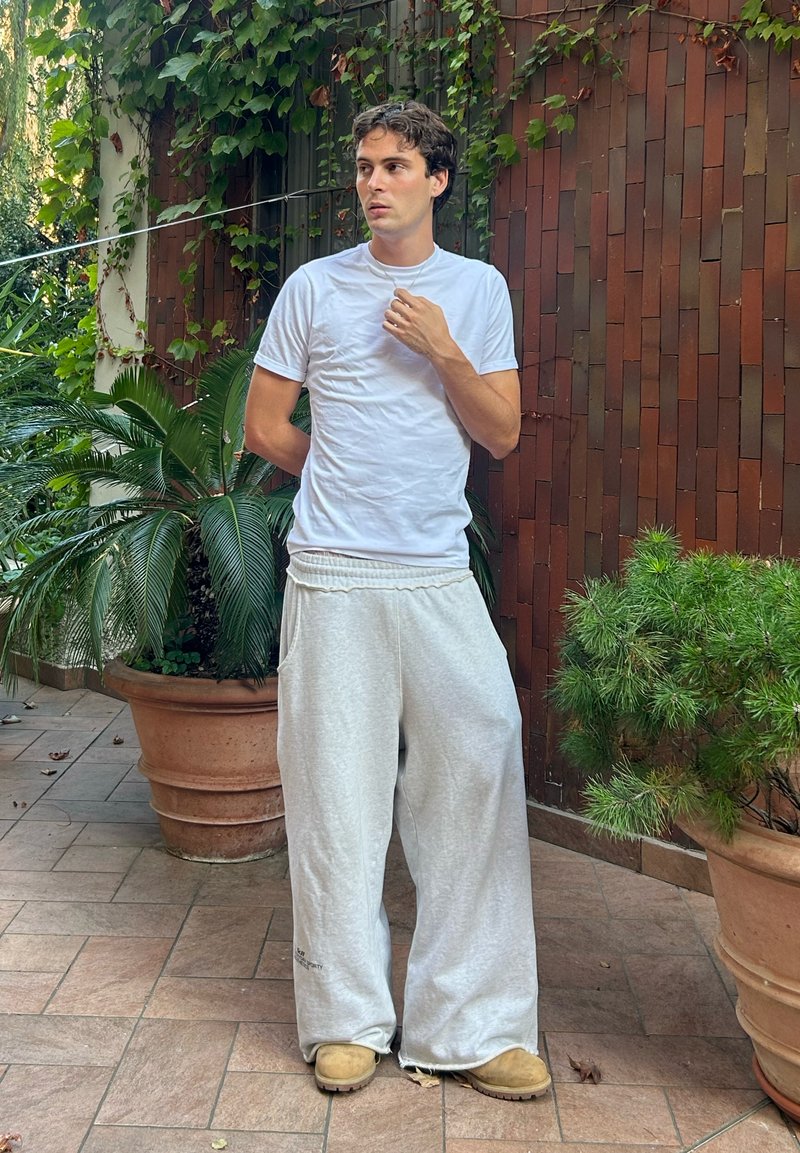 Weisses T-Shirt mit kurzen Ärmeln, hellgraue oversized Sweatpants und bequeme Slip-On-Schuhe in Beige. Im Hintergrund grüne Pflanzen und eine strukturiert braune Wand.