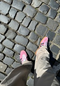 Růžové semišové tenisky s černými pruhy a bílými tkaničkami, nošené s béžovými kalhotami, na strukturovaném dlažebním povrchu.