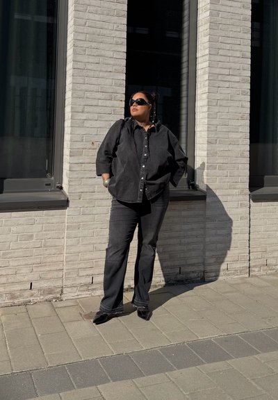 Camisa de mezclilla negra de botones combinada con pantalones de mezclilla negra y zapatos negros brillantes. El escenario presenta un fondo urbano iluminado por el sol.