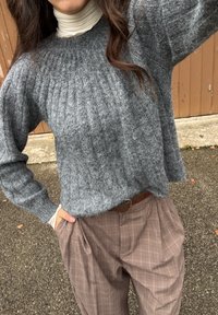 Femme portant un pull en maille gris sur un col roulé blanc et un pantalon à carreaux marron avec ceinture, debout en plein air avec une main dans la poche.