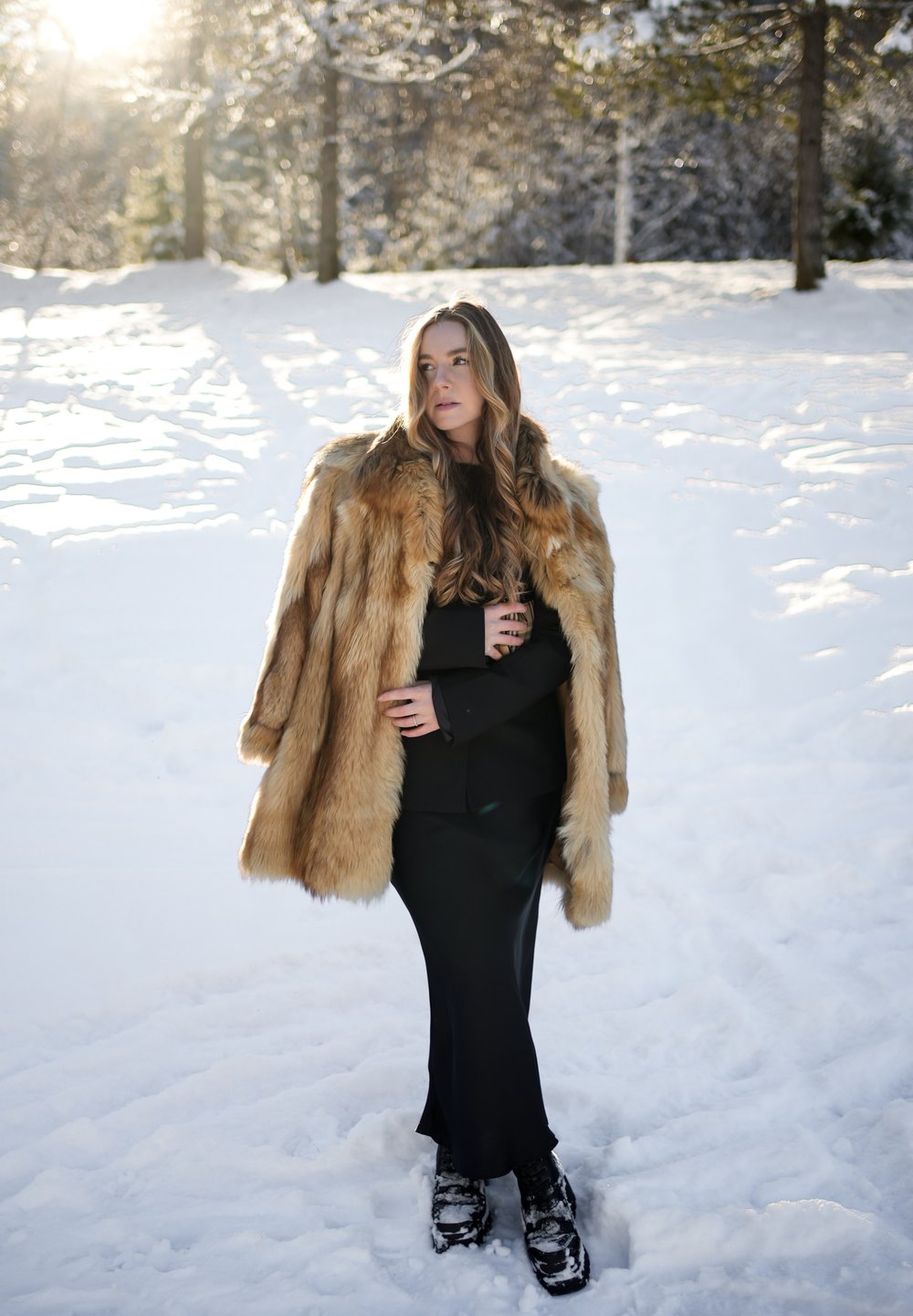 Femme aux cheveux longs portant un manteau de fourrure par-dessus une robe noire, se tenant sur la neige, avec la lumière du soleil filtrant à travers des arbres couverts de gel en arrière-plan.