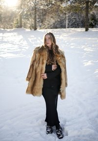 Femme aux cheveux longs portant un manteau de fourrure par-dessus une robe noire, se tenant sur la neige, avec la lumière du soleil filtrant à travers des arbres couverts de gel en arrière-plan.
