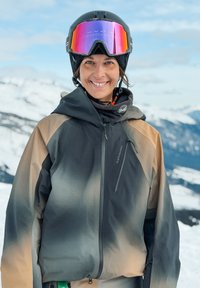 Χαμογελαστό άτομο που φοράει μπεζ και γκρι μπουφάν για σκι, μαύρο κράνος με ροζ αντανακλαστικά γυαλιά, στέκεται σε χιονισμένο ορεινό τοπίο.