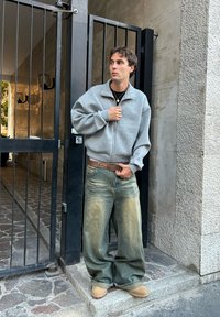 Sudadera gris con cremallera, jeans azules desgastados y holgados, cinturón marrón, zapatos de lona color beige, de pie junto a una puerta con fondo de muro de piedra.