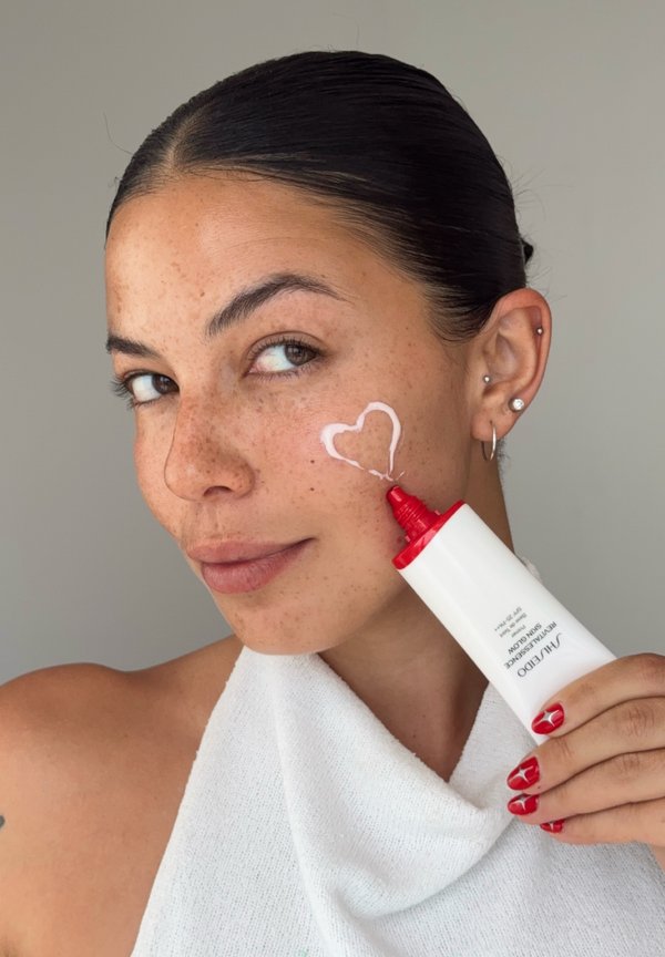 Femme aux cheveux foncés et taches de rousseur applique une crème blanche en forme de cœur sur sa joue en utilisant un tube à embout rouge, portant un haut en serviette blanche.