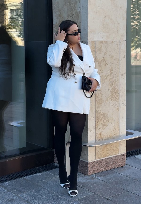 Mujer con cabello largo y oscuro, vestida con una blazer blanca de doble botonadura, medias negras, tacones blancos abiertos en la punta, gafas de sol negras, y sosteniendo un bolso negro.