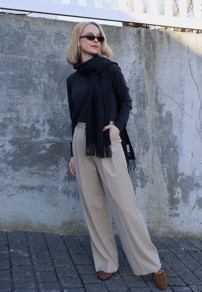 Mujer con gafas de sol, suéter negro y bufanda, pantalones beige de pierna ancha y zapatos marrones, de pie frente a una pared gris con textura.