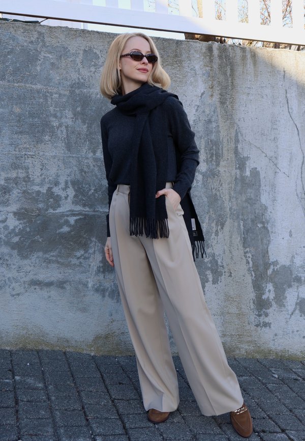 Frau mit Sonnenbrille, schwarzem Pullover und Schal, beigen Hosen mit weitem Bein und braunen Schuhen, die vor einer strukturierten grauen Wand steht.