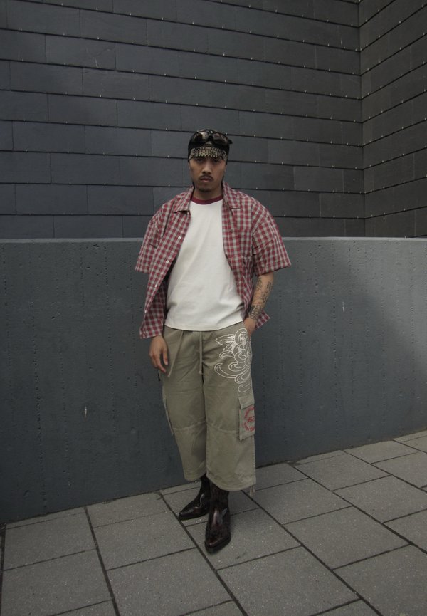 Homme portant une chemise à carreaux rouge sur un t-shirt blanc, pantalon beige large et court avec des motifs, bottes noires, bandana et lunettes de soleil, debout dans un coin gris.