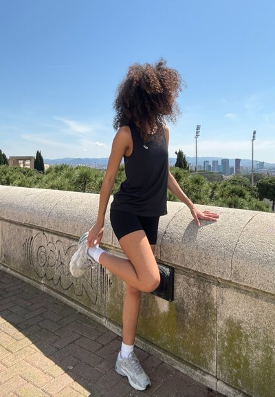 Top negro sin mangas de estilo deportivo combinado con pantalones cortos negros. La persona lleva calcetines blancos y zapatillas grises, de pie frente a una pared de piedra.