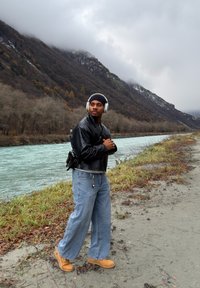 Veste en cuir noire, chemise gris clair, jeans larges bleus, bottes beige et écouteurs argentés. Debout près d'une rivière turquoise, avec des montagnes en arrière-plan.