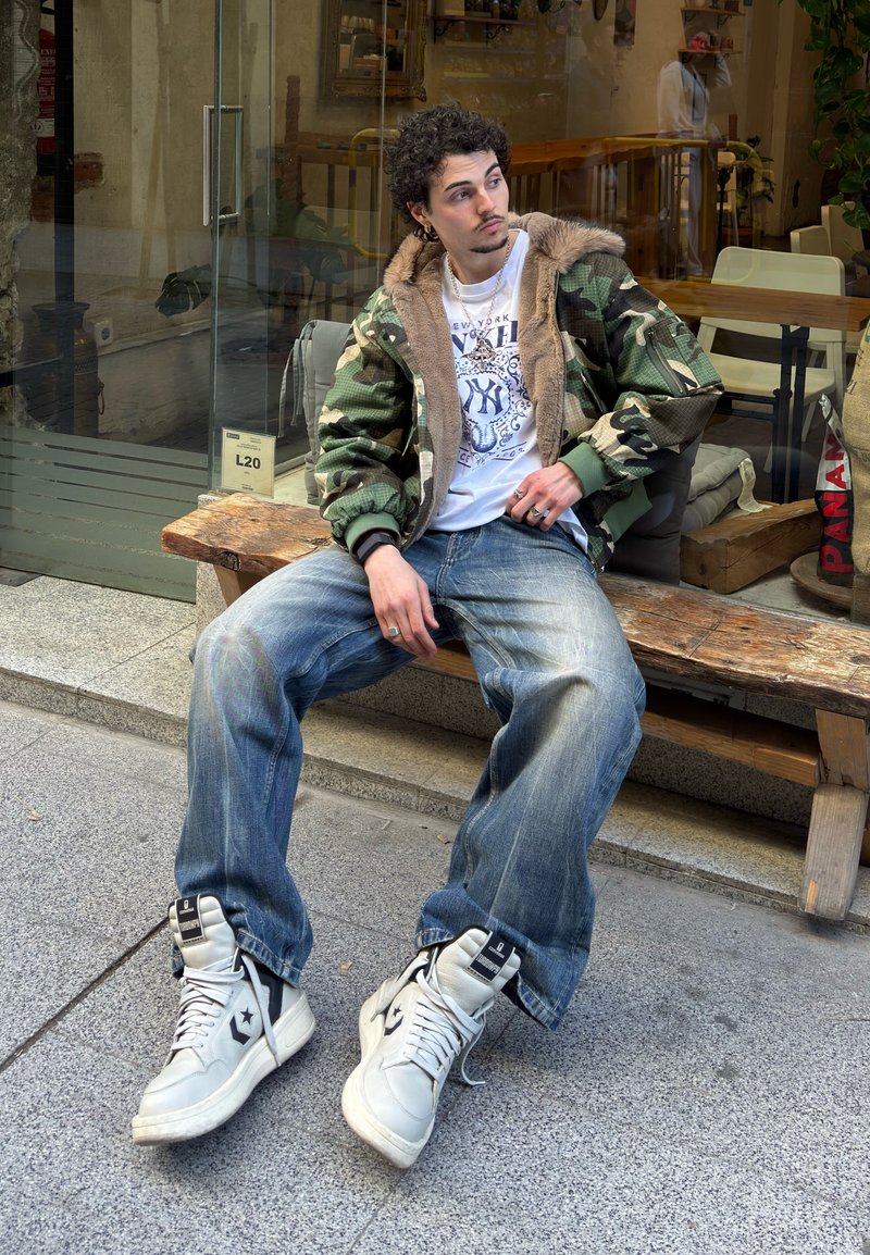 Jeune homme aux cheveux bouclés portant une veste camouflage et des baskets, assis détendu sur un banc en bois devant un café vitré.