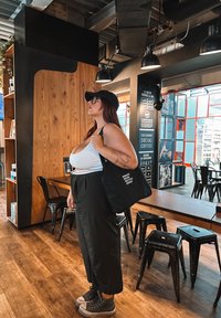 Saco tote preto com texto branco, camisola branca folgada, calças pretas de cintura alta e ténis cinzentos num ambiente de café com decoração em madeira e metal.