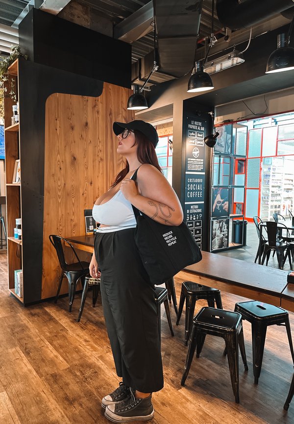 Borsa a tracolla nera con testo bianco, canotta bianca larga, pantaloni neri a vita alta e sneaker grigie in un caffè arredato con elementi in legno e metallo.