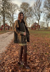 Hnědá voskovaná bunda s kostkovanou podšívkou přes zelené pletené šaty. Tmavě hnědé kozačky až ke kolenům. Zem pokrývají podzimní listy.