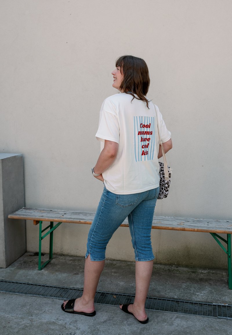 Femme portant un T-shirt blanc avec le texte "Cool mums have cool kids" dans le dos, pantalon capri en denim, sandales noires, debout près d'un banc en bois.