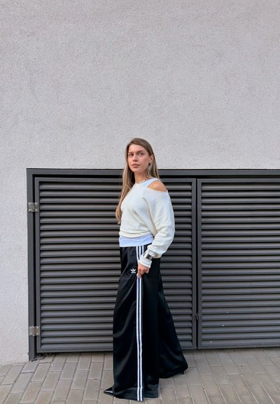 Mujer joven con el pelo largo que lleva un suéter blanco con los hombros descubiertos y pantalones anchos negros con rayas blancas, está junto a una pared gris con rejilla de ventilación.