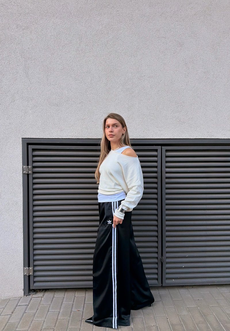 Jeune femme aux longs cheveux portant un pull blanc à épaules dénudées et un pantalon large noir avec des bandes blanches, debout près d'un mur gris ventilé.