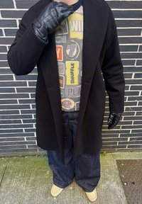 Személy fekete kabátot, mintás pulóvert, fekete bőrkesztyűt, széles kék farmert és bézs cipőt visel, sötét téglafal előtt áll.