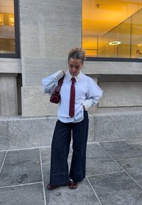 Camicia a righe bianche e blu con maniche oversize, abbinata a pantaloni in denim scuro a gambe larghe. Accessori includono una cravatta bordeaux e una piccola borsa.