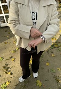 Beige trenssitakki kermaisen logollisen villapaidan päällä, mustat housut ja valkoiset lenkkarit. Kädessä mustat aurinkolasit ja ranteessa koristeellinen rannekoru.