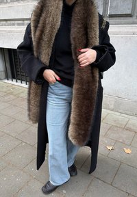 Personne portant un manteau noir avec un large col en fourrure marron, un pull noir, un jean bleu clair et des chaussures noires, debout sur un trottoir en ville.