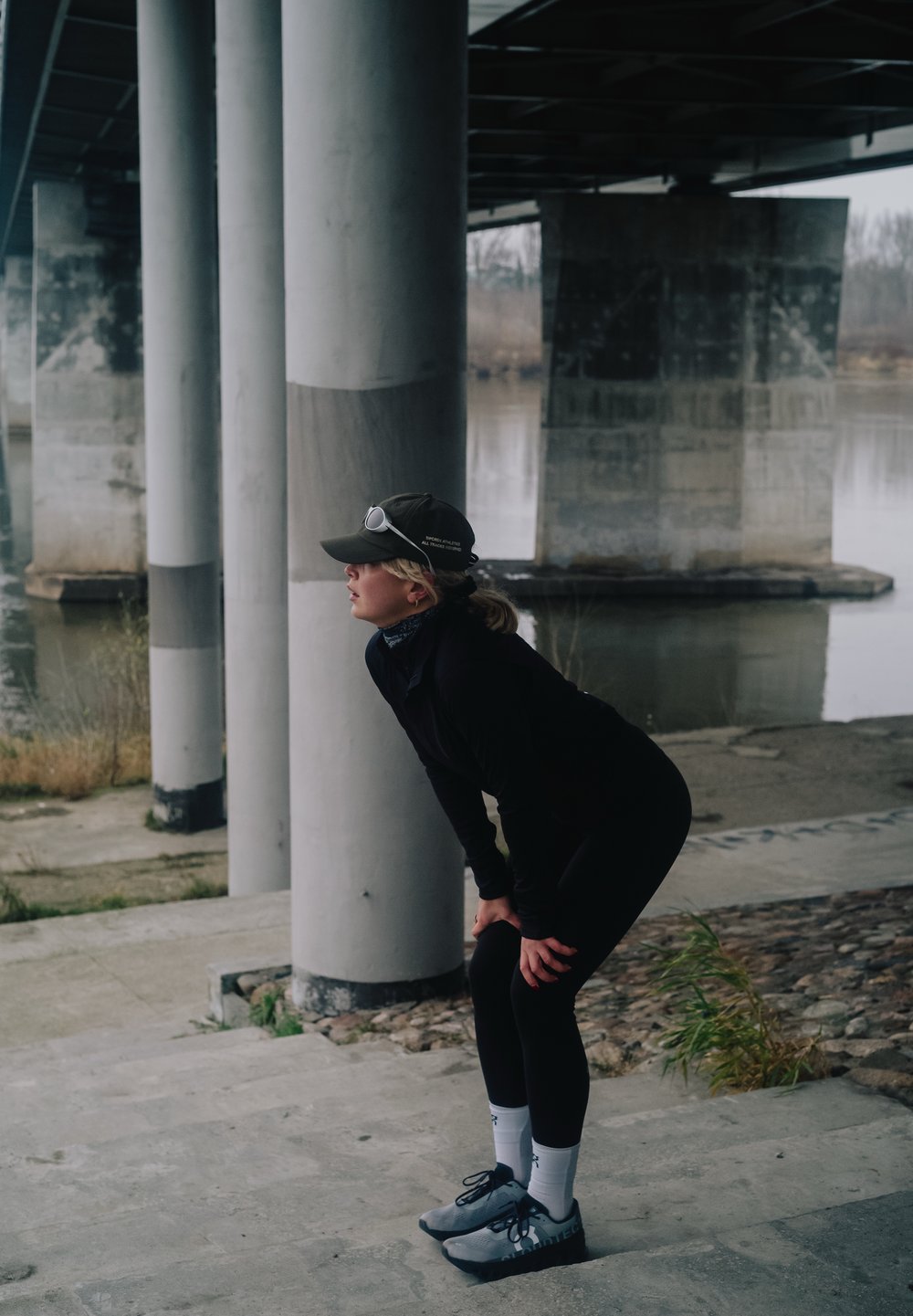 Persoon in zwarte sportkleding en pet die voorover buigt en met handen op knieën rust onder een betonnen brug bij een waterlichaam.