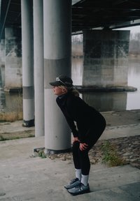 Persoon in zwarte sportkleding en pet die voorover buigt en met handen op knieën rust onder een betonnen brug bij een waterlichaam.