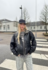 Blouson aviateur en cuir noir avec fermeture éclair et poches, associé à un sweat-shirt graphique gris et un pantalon large imprimé serpent, sous une casquette noire.