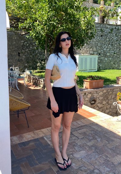 Camisa polo blanca de manga corta, falda mini negra acampanada, bolso de paja beige tejido, gafas de sol oscuras y chanclas negras contra un fondo de pared de piedra.