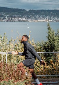 Man in black sportswear jogging outdoors near a lake, with a hillside town visible across the water in the background.