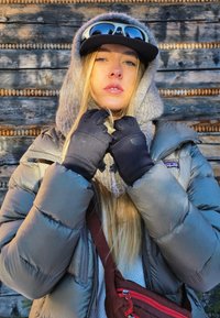 Opgeblazen grijze jas, gebreide grijze capuchon, zwarte handschoenen en zwarte pet met zonnebril. Het model heeft lang blond haar tegen een houten achtergrond.