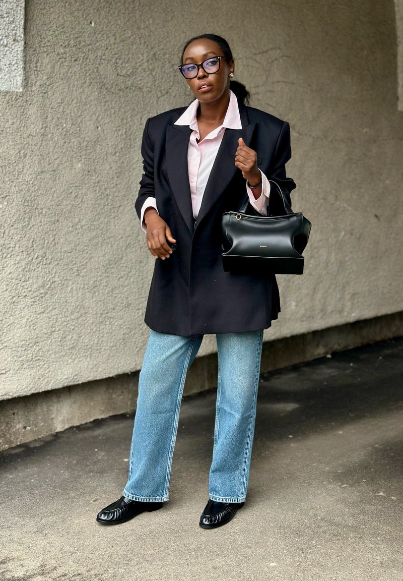 Žena v černém oversized saku, světle růžové košili, modrých džínách, černých mokasínách a brýlích, nesoucí černou kabelku, stojící opřená o texturovanou zeď.