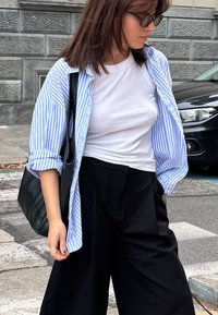 Mulher vestindo calças pretas, camisa branca, camisa aberta com riscas azuis, óculos de sol, a carregar uma bolsa preta, em pé numa rua da cidade perto de uma passadeira.