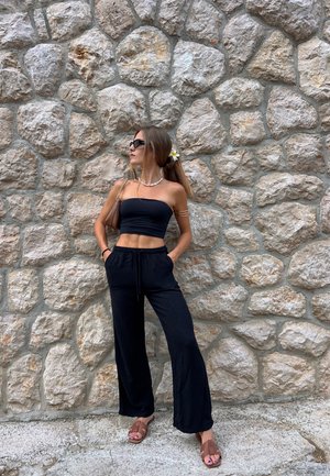 Top corto negro tipo tubo y pantalones negros holgados. Sandalias marrones con un diseño de tiras en cruz. Flor blanca y amarilla en el cabello. Fondo de pared de piedra.