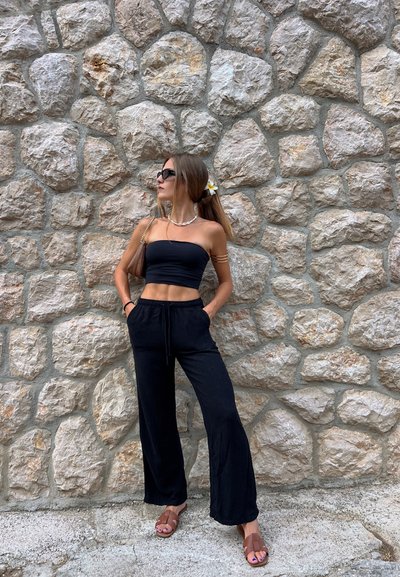 Top corto negro tipo tubo y pantalones negros holgados. Sandalias marrones con un diseño de tiras en cruz. Flor blanca y amarilla en el cabello. Fondo de pared de piedra.