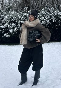 Veste rembourrée marron à carreaux avec un col en fourrure, pantalon noir surdimensionné, bottes en caoutchouc grises et sac à main noir, sur fond enneigé.