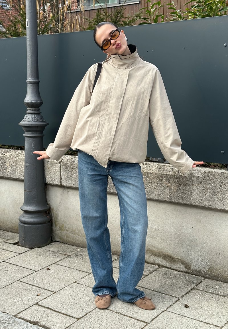Mujer con chaqueta beige, vaqueros azules, zapatos planos marrones y gafas de sol posando al aire libre cerca de un farol y una pared de hormigón.