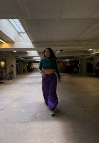 Una persona lleva una camiseta de manga larga verde corta con rayas blancas y pantalones de cargo morados sueltos, caminando en un garaje de estacionamiento.