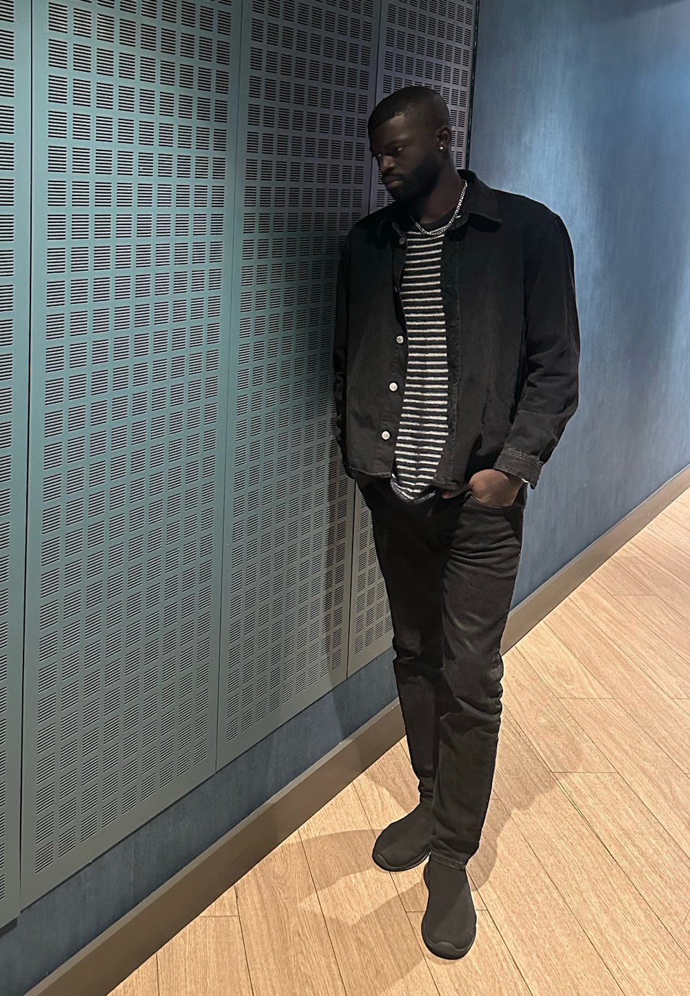 Άντρας με γενειάδα και σκουλαρίκια, που φοράει μαύρο σακάκι και ριγέ πουκάμισο, στέκεται με τα χέρια στις τσέπες δίπλα σε μπλε διάτρητο τοίχο και ξύλινο πάτωμα.