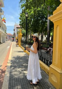 Model in a sleeveless white maxi dress with a ruffled hem and a patterned headband, holding a drink, on a tree-lined street.
